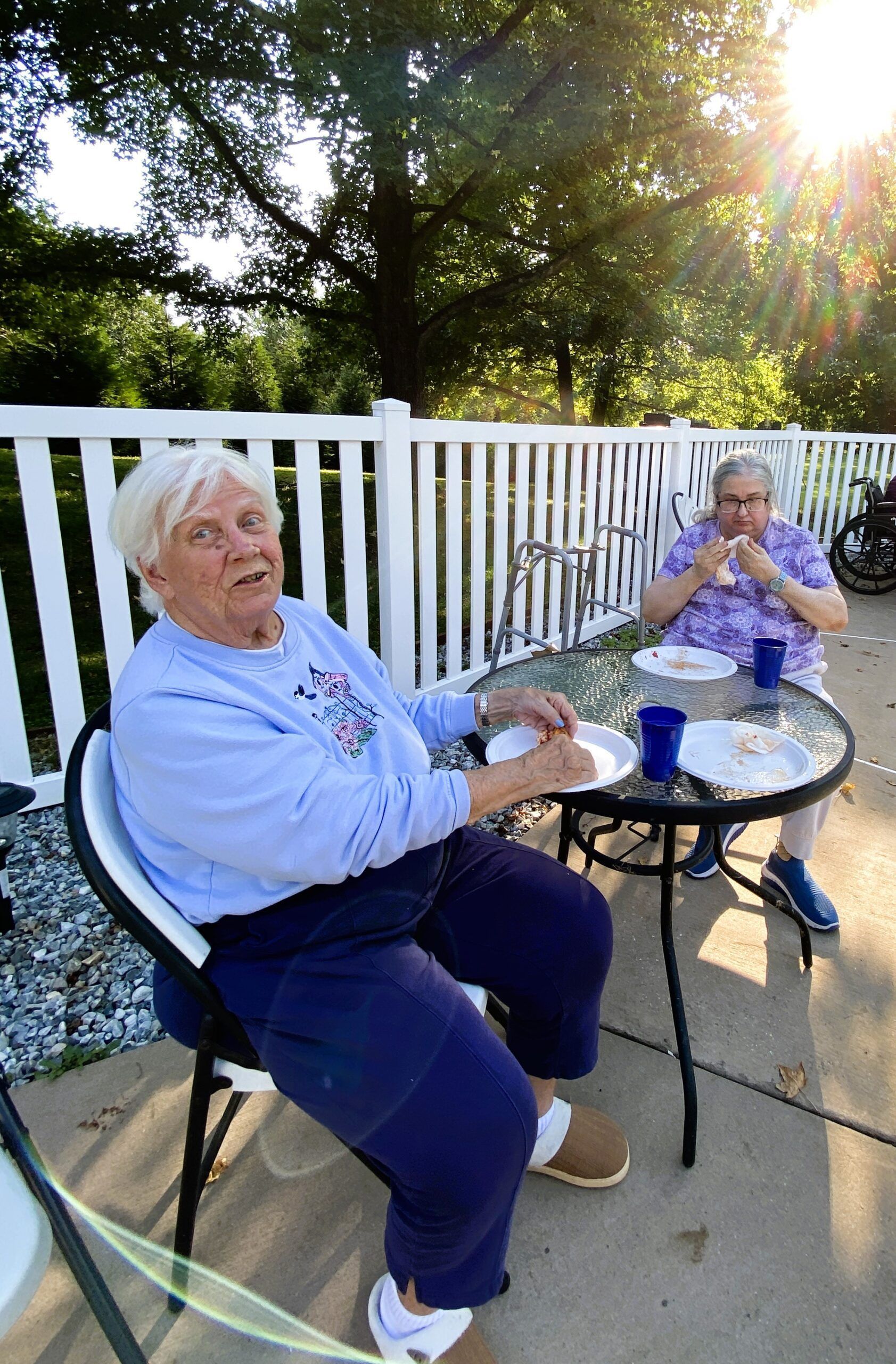 Two people sit at a patio table outdoors eating, with one person facing the camera in the foreground.