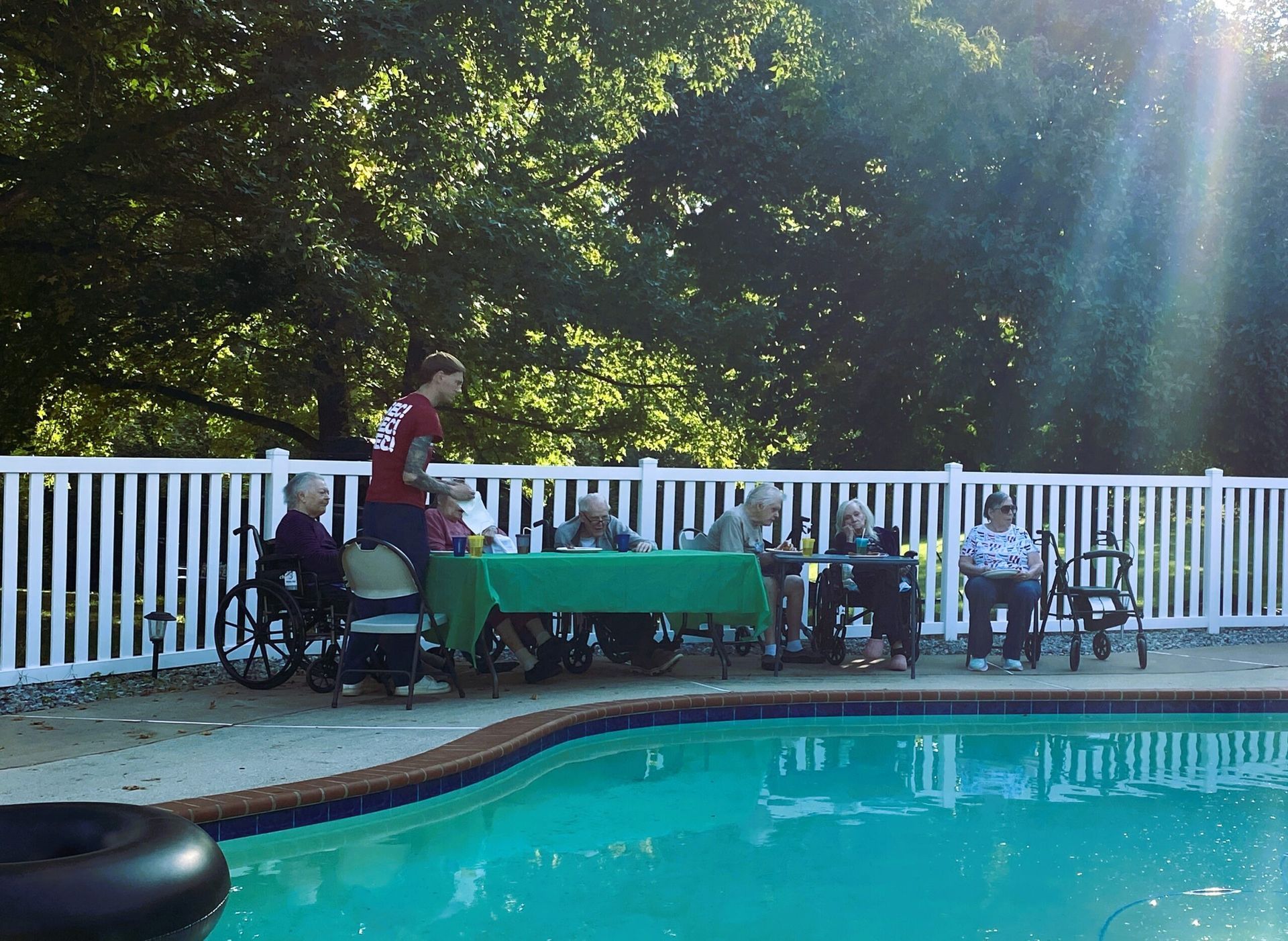 A group of people sit at a green-covered table by a swimming pool behind a white fence under a sunlit tree canopy.