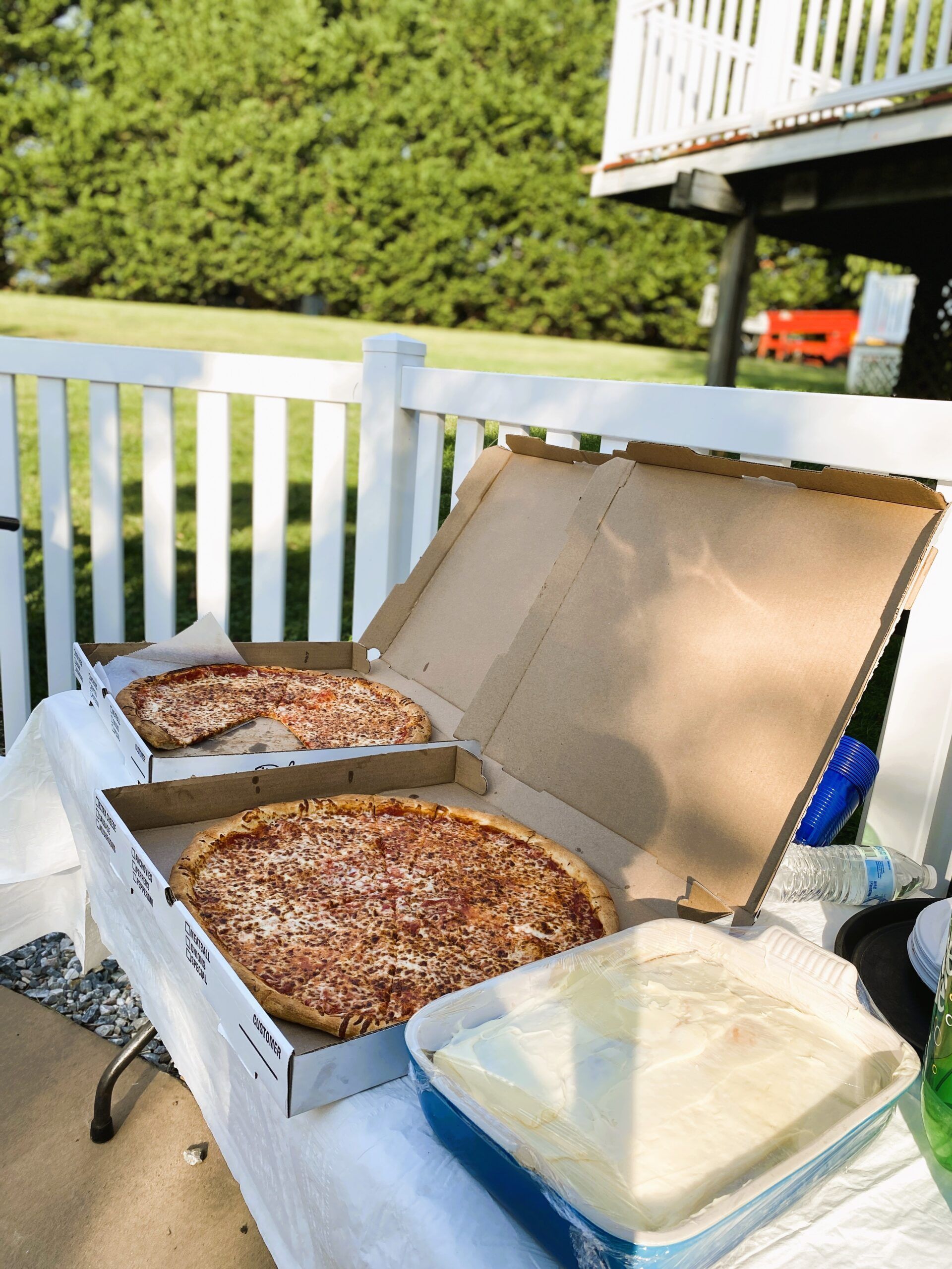 Two open pizza boxes and a dish of white food sitting on a table outside next to a white fence.
