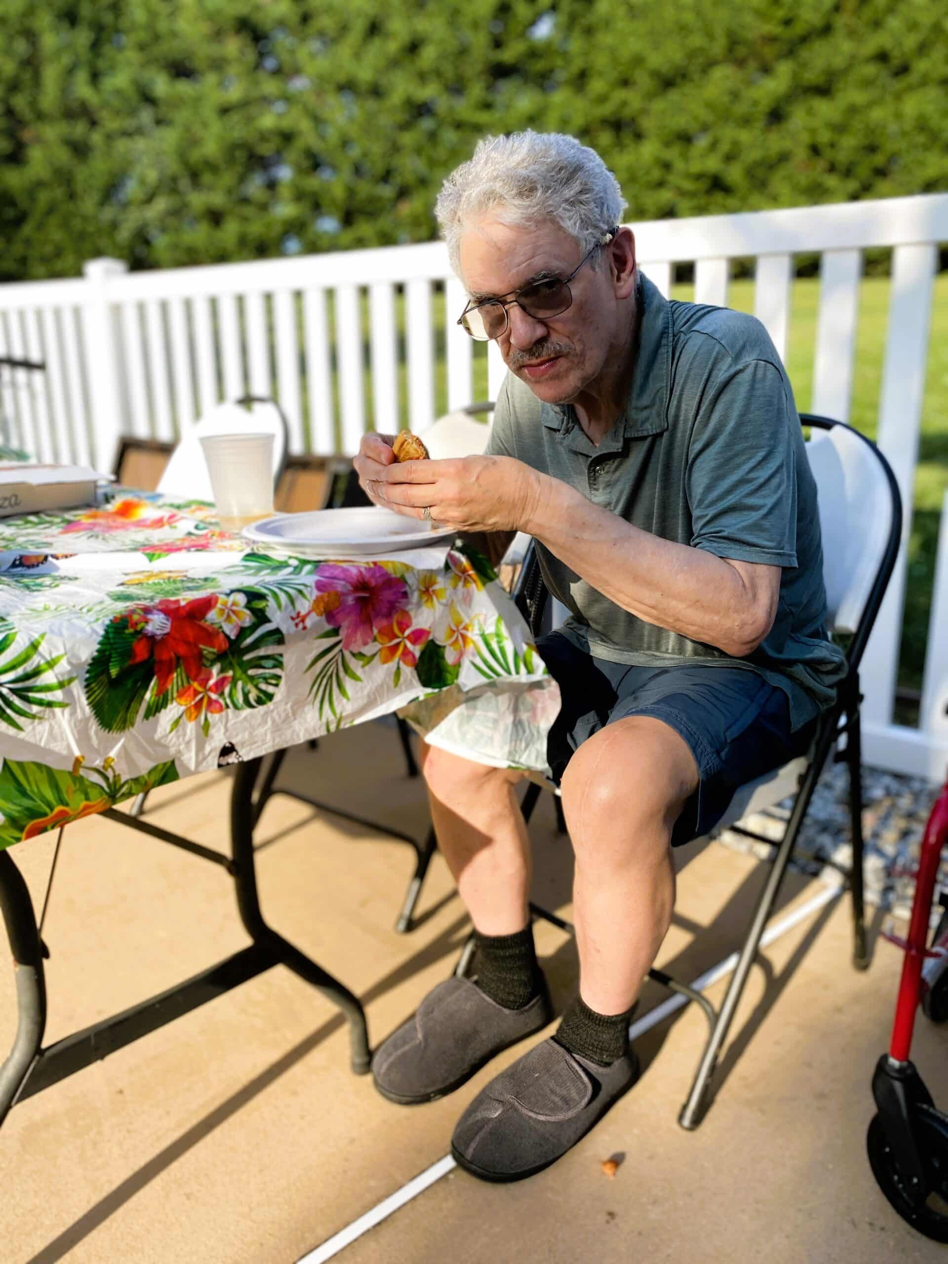 A person sits at an outdoor table with a floral tablecloth, eating a snack while wearing sunglasses and dark slippers.