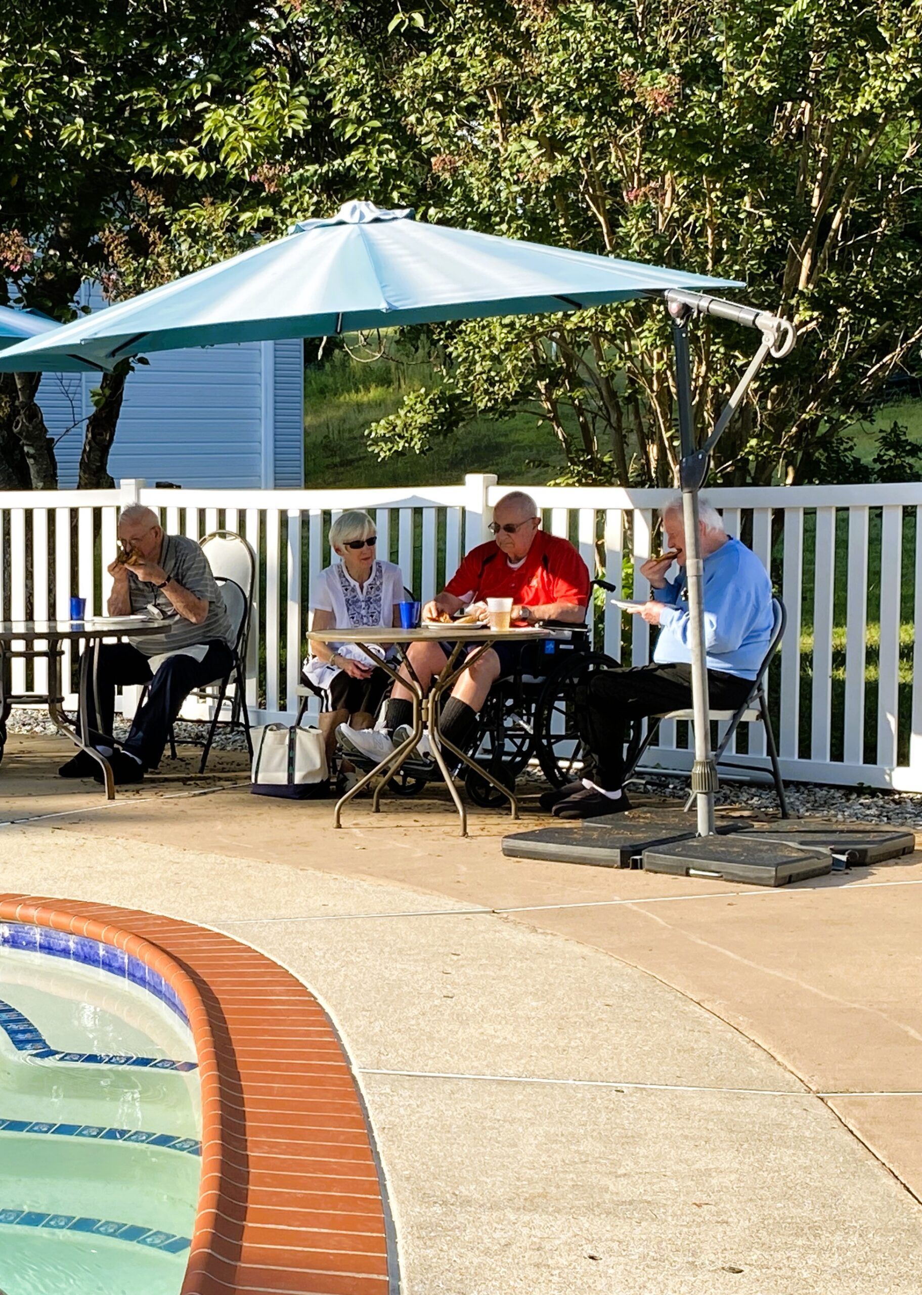 Four people sit at two outdoor tables under a blue umbrella by a swimming pool, eating and talking.