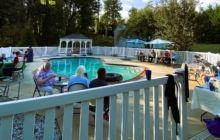 A poolside outdoor gathering with people seated at tables near a gazebo, viewed from behind a white fence.