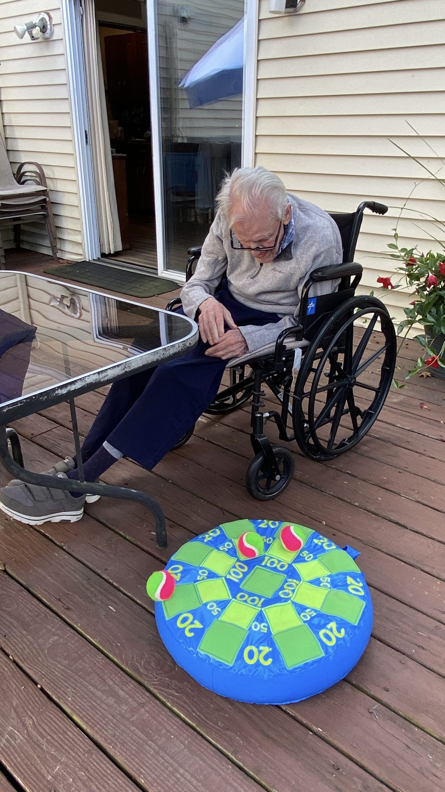 An individual in a wheelchair sits on a deck near a blue and green inflatable dartboard target on the floor.