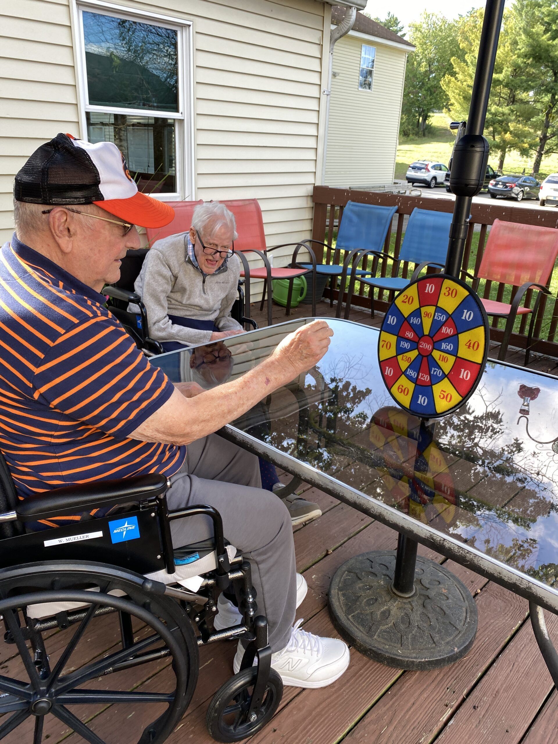 Two people in wheelchairs play a dart game on a patio table outside a house.