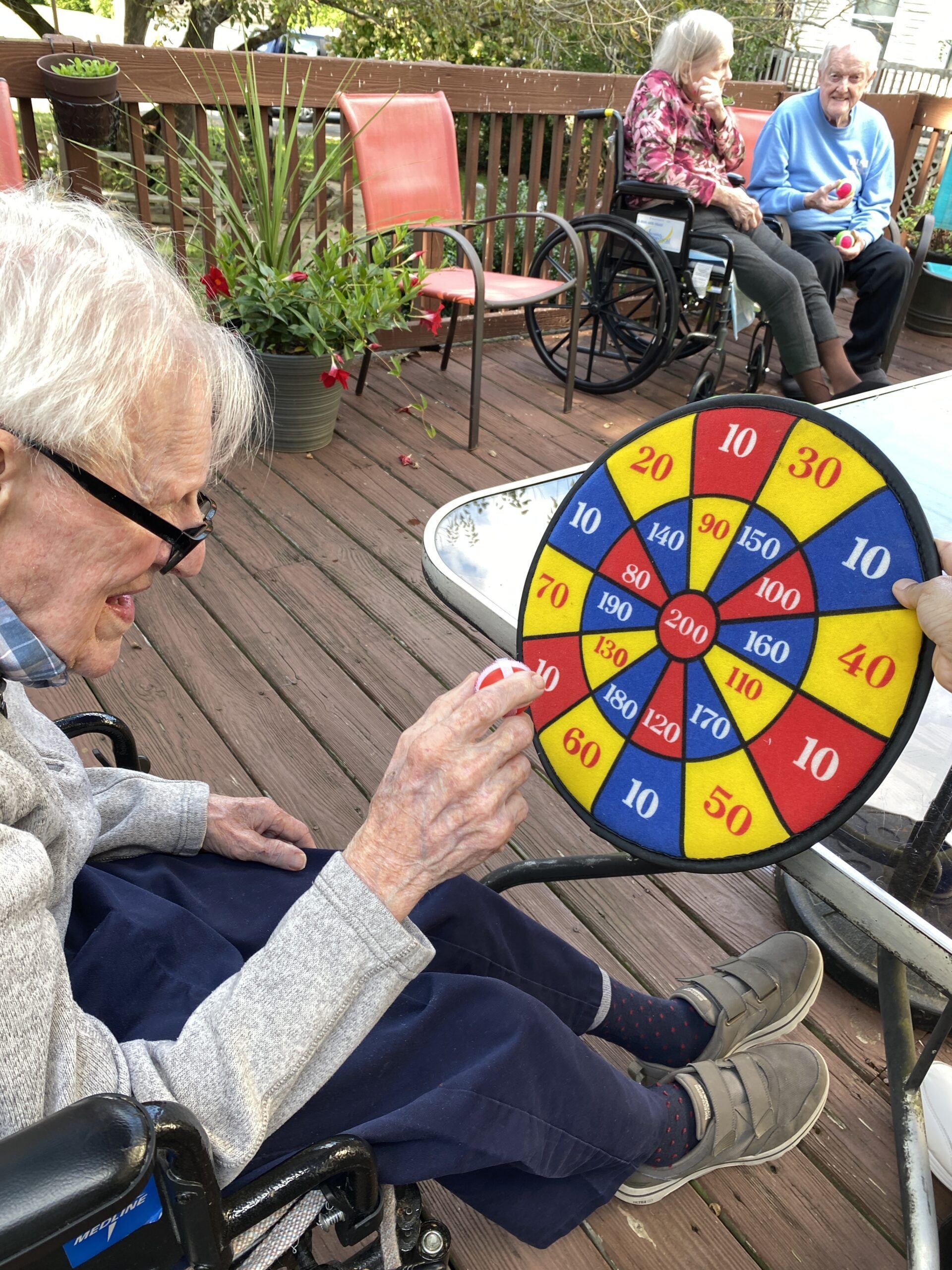 People in a sunny outdoor setting play with a colorful target board held up by a hand.