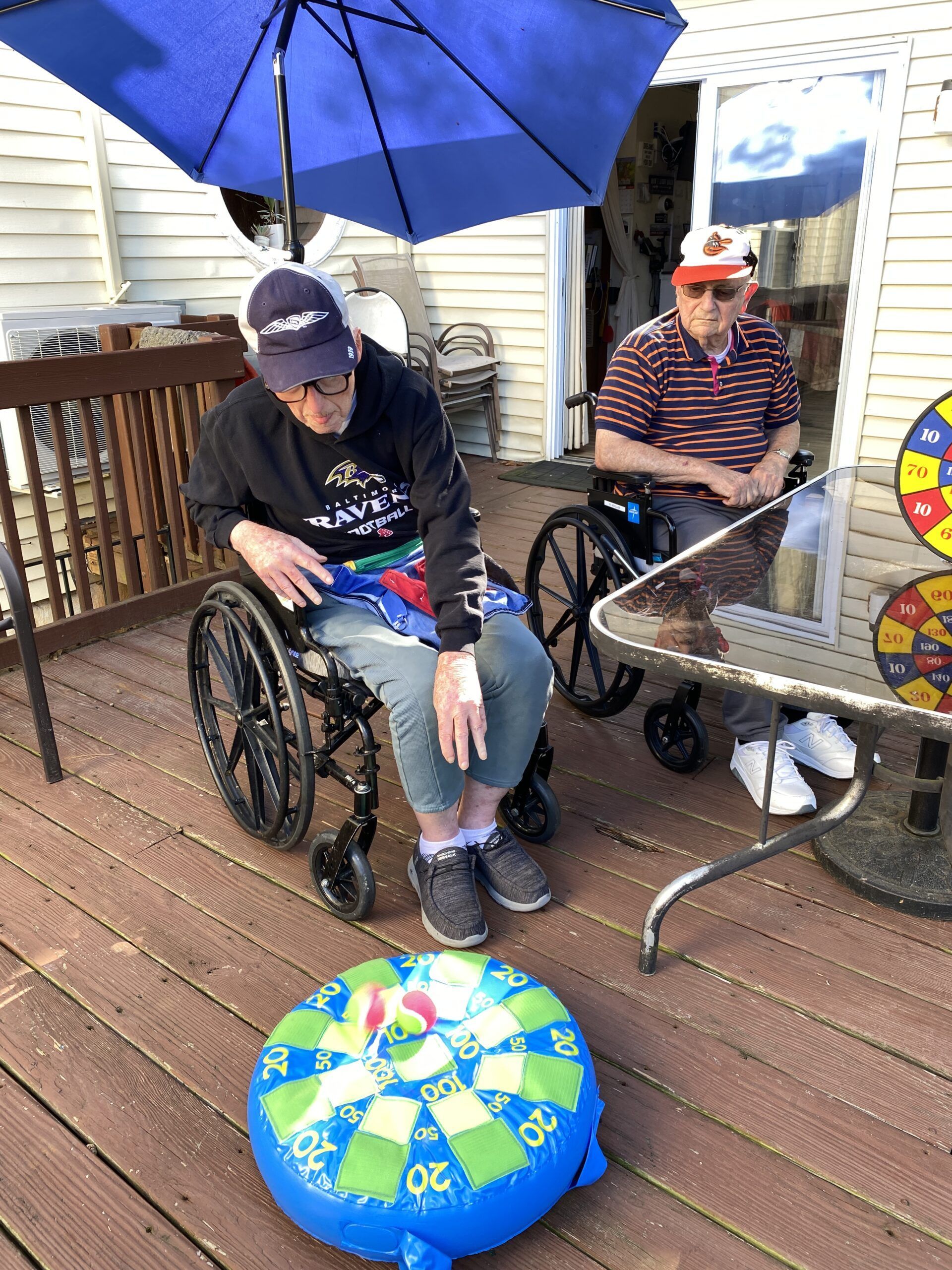 Two people in wheelchairs play a floor dart game on a wooden deck under a blue umbrella.