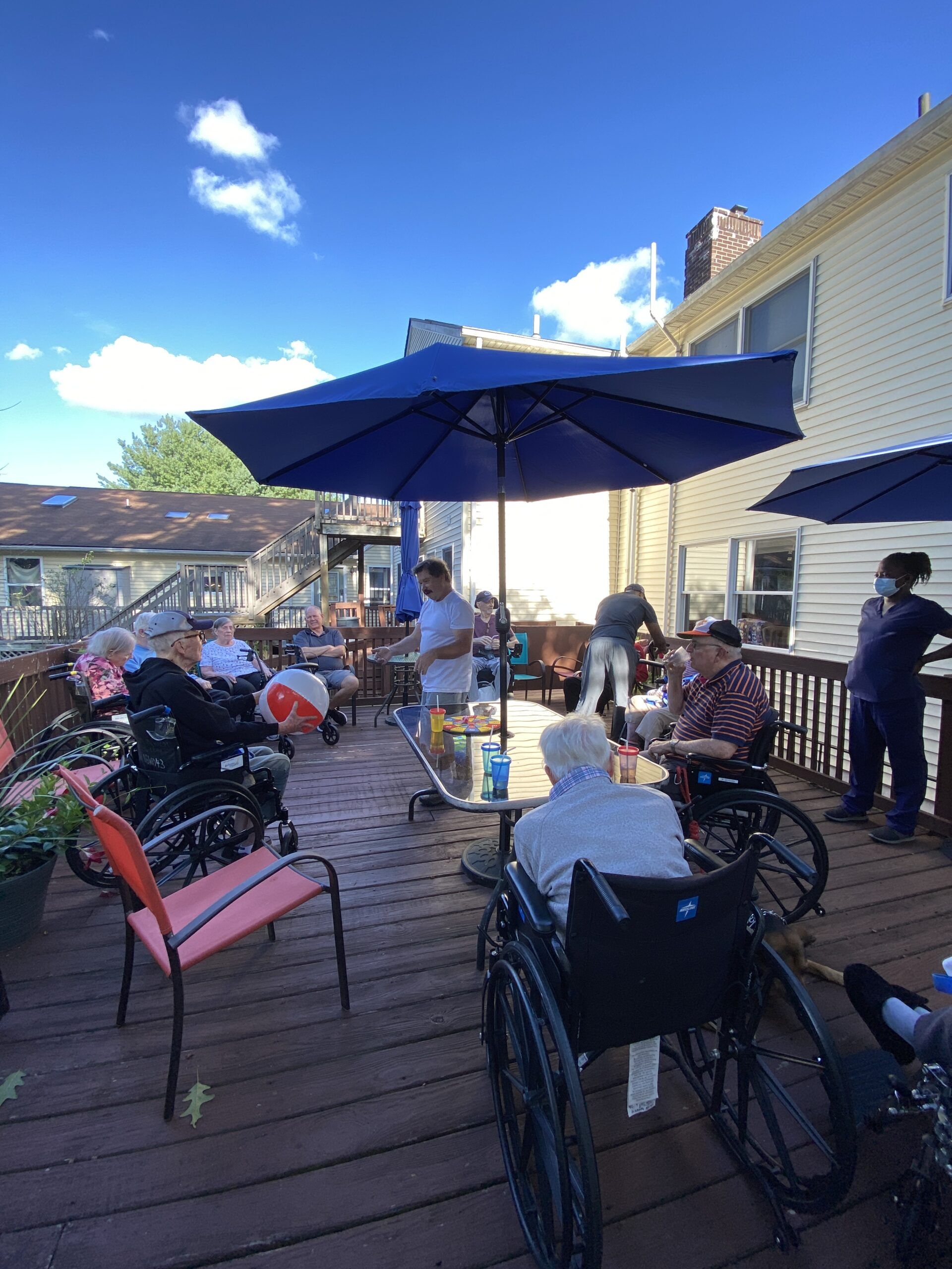People in wheelchairs participate in a group activity on a sunlit wooden deck under a large blue umbrella.
