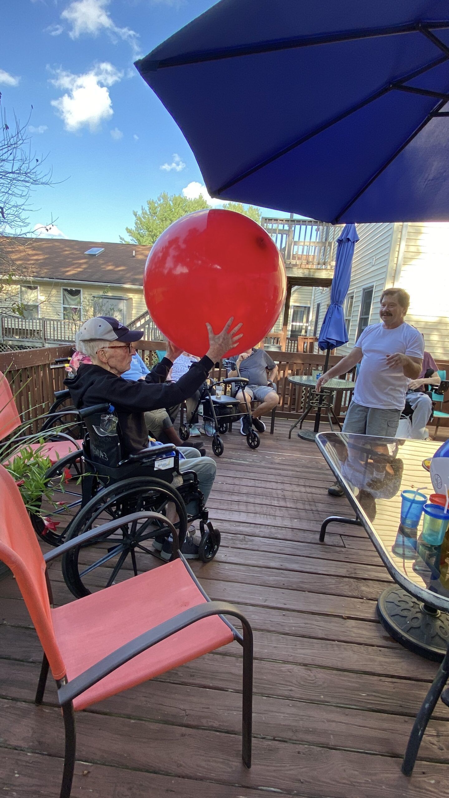 A person in a wheelchair holds a large red ball on a deck, with others nearby under a blue patio umbrella.