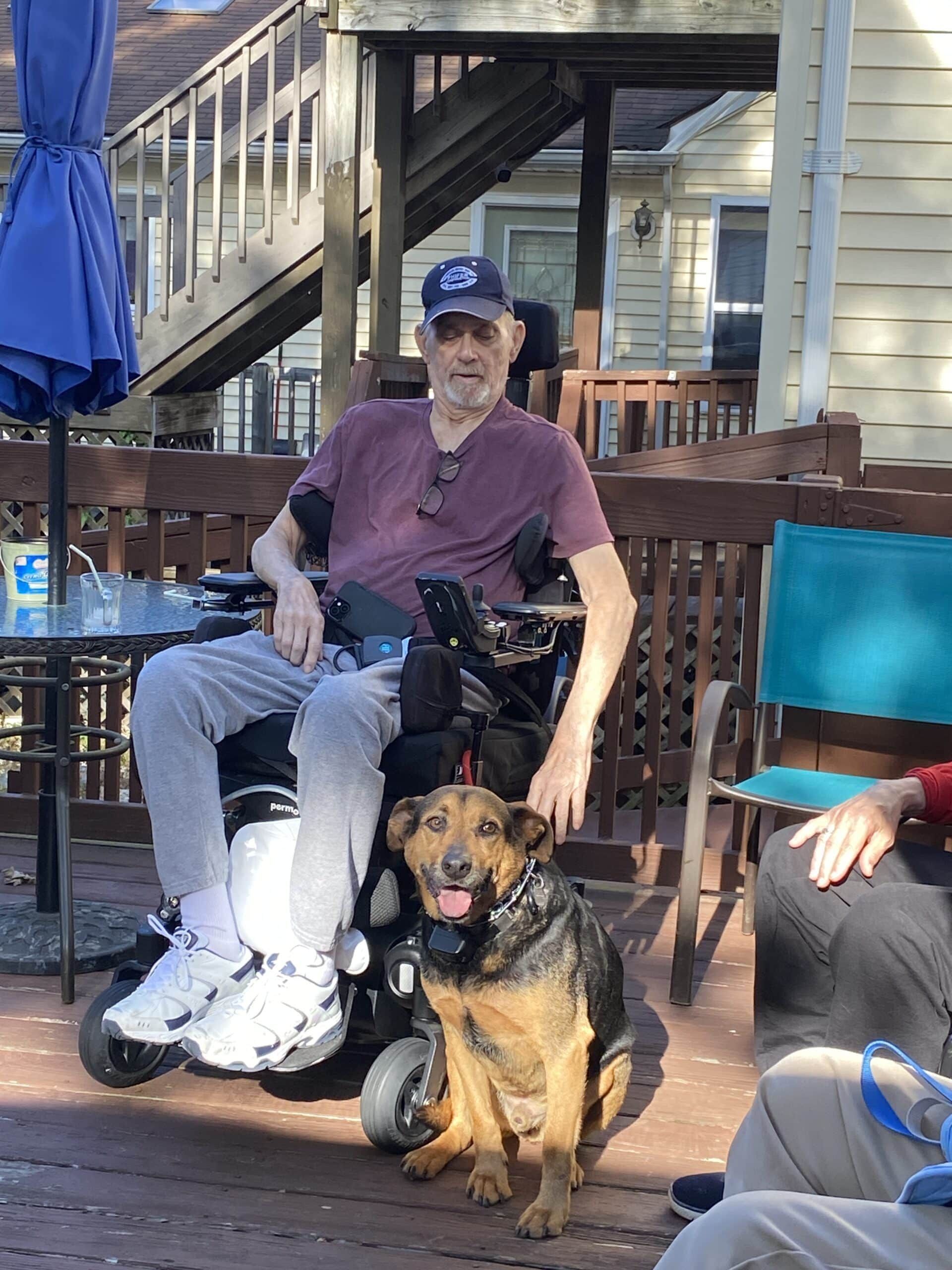 A person in a wheelchair sits on a wooden deck next to a smiling dog, with a table and outdoor furniture nearby.