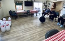 People participating in an indoor bowling activity, with one person rolling a black ball toward a set of white pins.