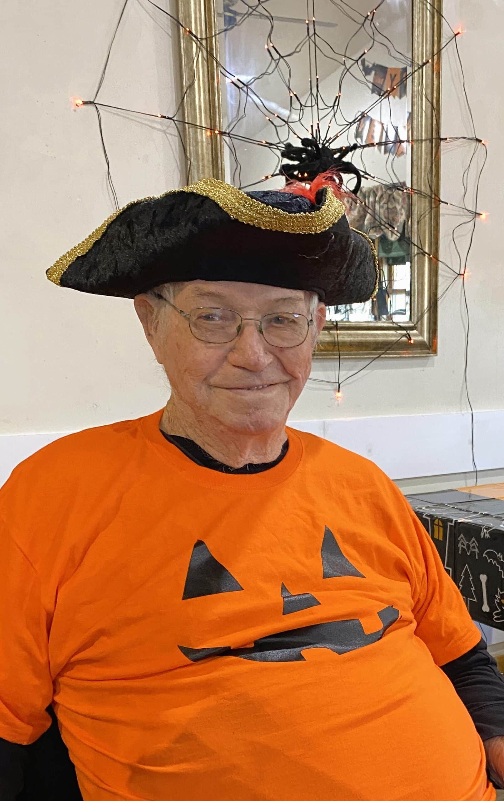 An elderly person wearing a pirate hat and an orange pumpkin-faced T-shirt, sitting in front of a spiderweb decoration.