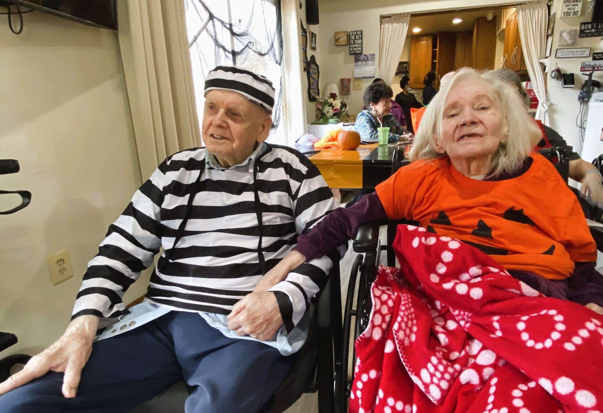 Two people in costumes, one as a prisoner and one in an orange shirt, sit together holding hands in an indoor space.