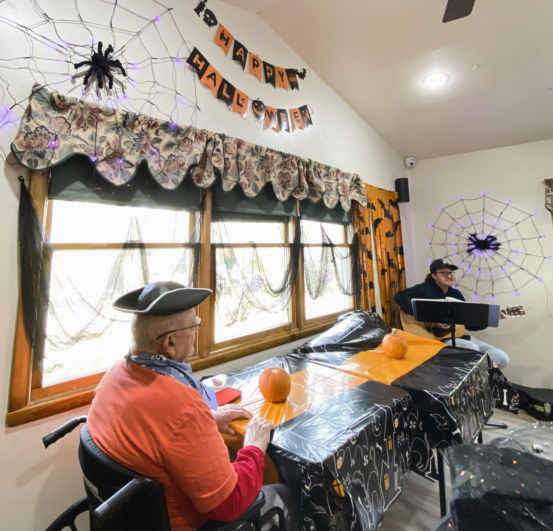 A senior in a pirate hat sits at a Halloween-decorated table while a musician plays guitar in a brightly lit room.