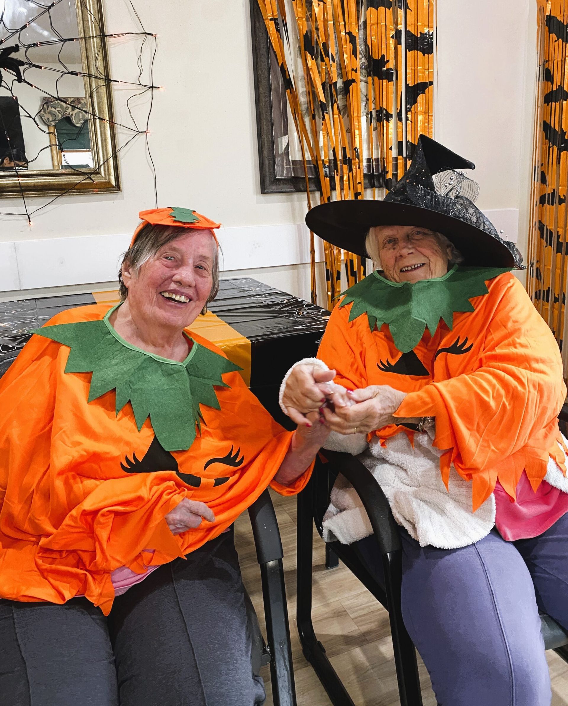 Two people in orange pumpkin costumes, one wearing a witch hat, sit together smiling at a Halloween-themed party.