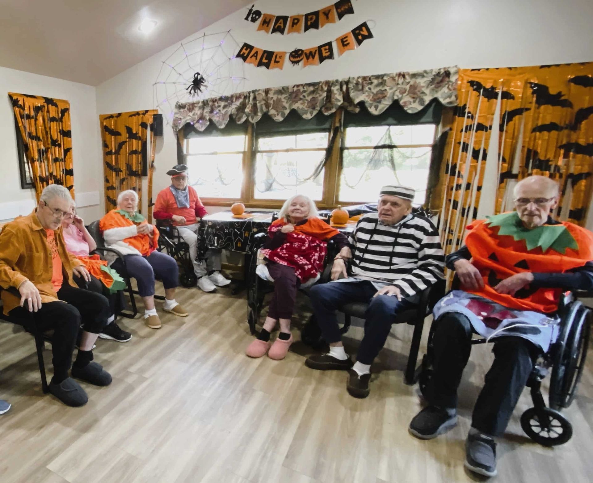 A group of people wearing Halloween costumes, including a pumpkin, gather in a festive room decorated for the holiday.