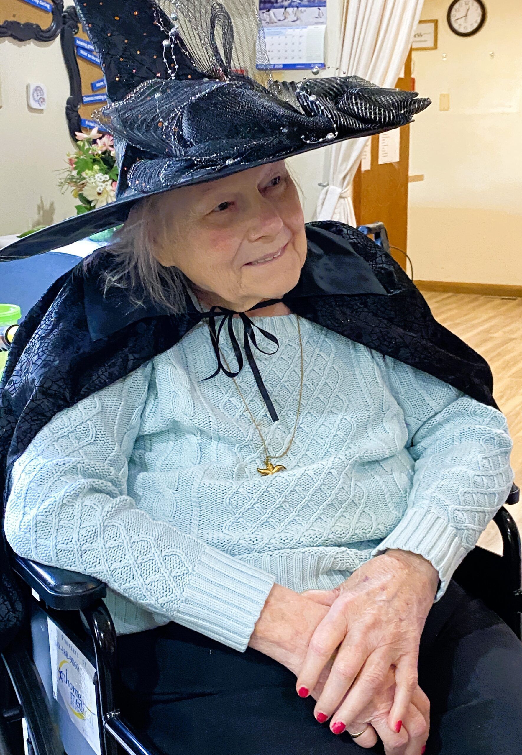 An individual in a wheelchair wears a black witch's hat and cape over a light blue sweater, smiling in an indoor setting.