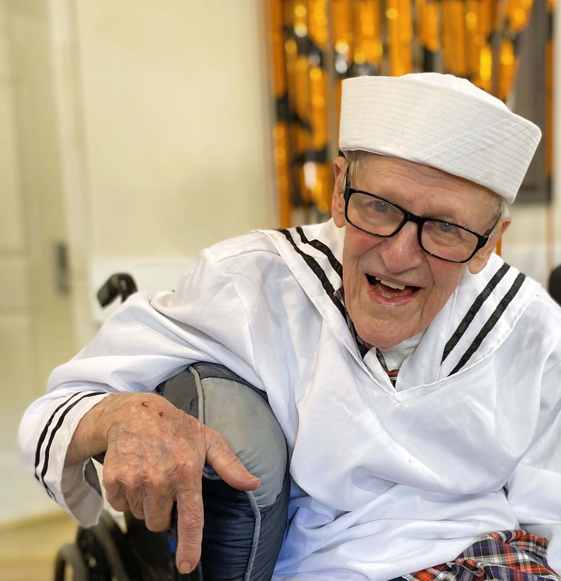 An elderly person in a white sailor uniform and cap smiles while sitting in a wheelchair.