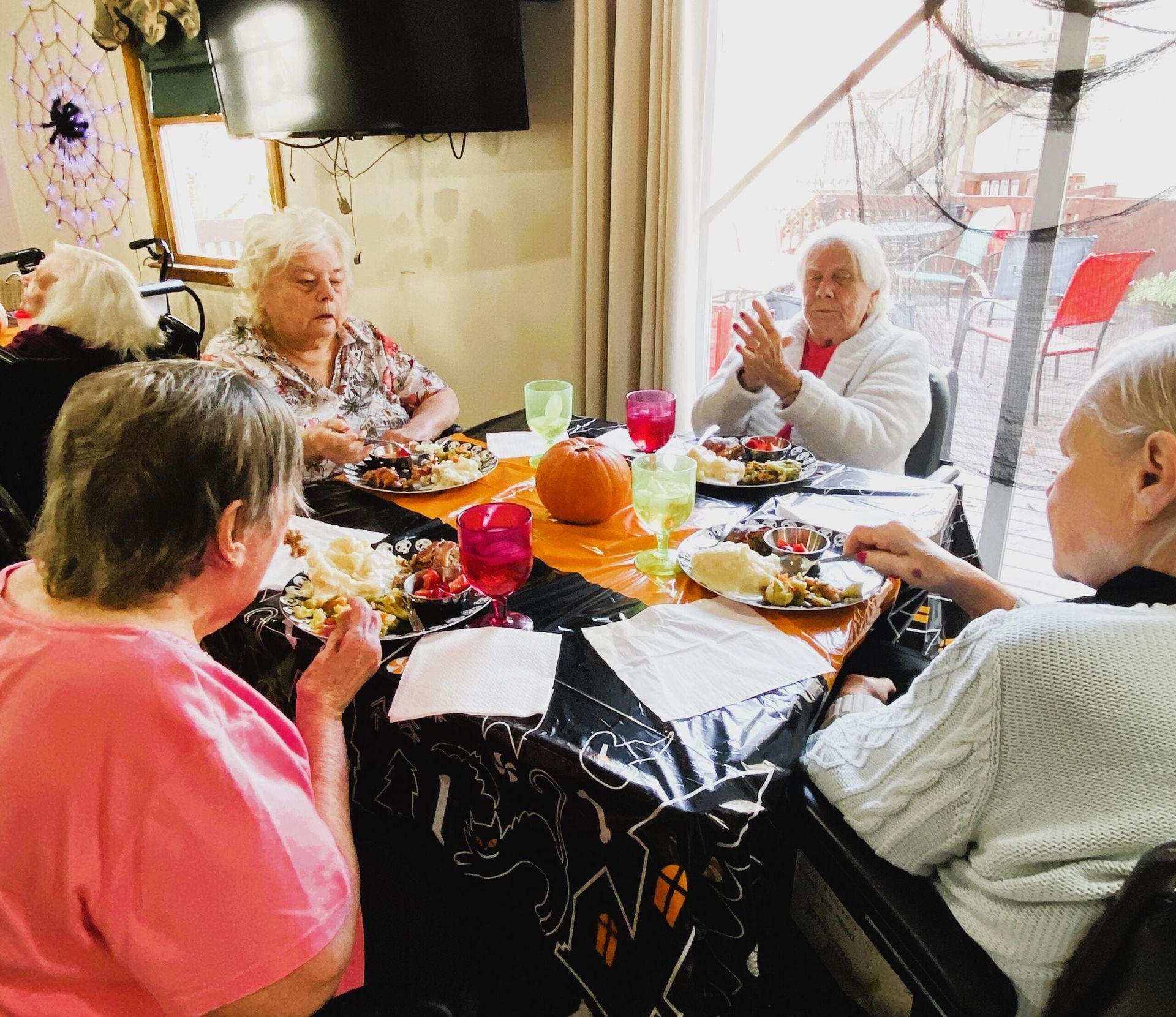 Four people sit around a table decorated with a Halloween tablecloth, eating a meal together in an indoor setting.