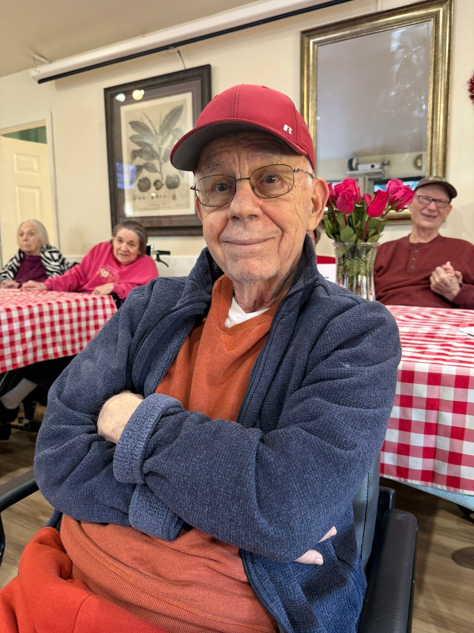 A person in a red cap and blue sweater sits with arms crossed in a dining room with others at tables with red-check cloths.