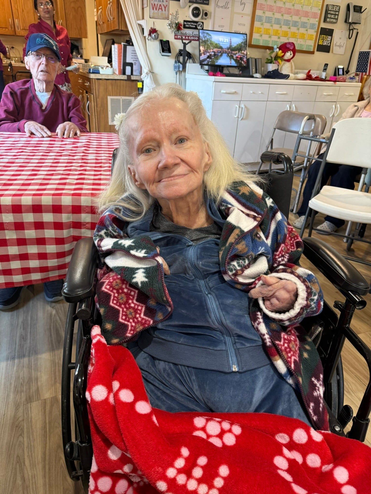 A person in a wheelchair with a red polka-dot blanket sits at a table in a room with a red-and-white checkered cloth.