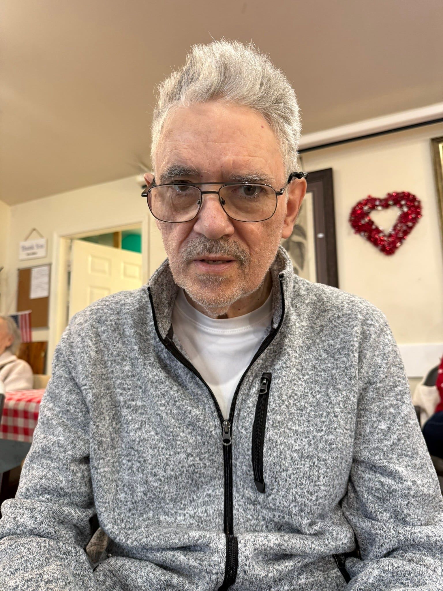 A person with short, light-colored hair and glasses wearing a gray zip-up fleece jacket, seated indoors.