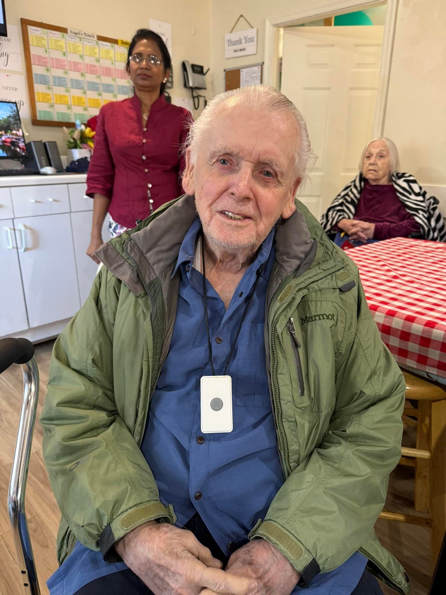An older individual sits in a room wearing a green jacket. Behind them, another person stands and one sits at a table.