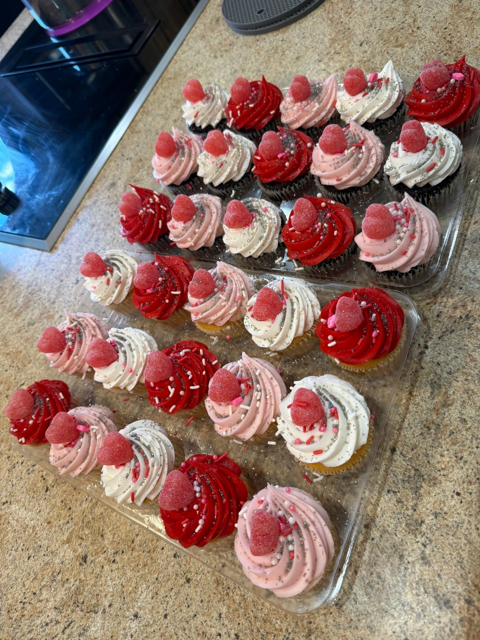 A tray of cupcakes decorated with red and pink frosting swirls, sprinkles, and a single gummy berry on each.