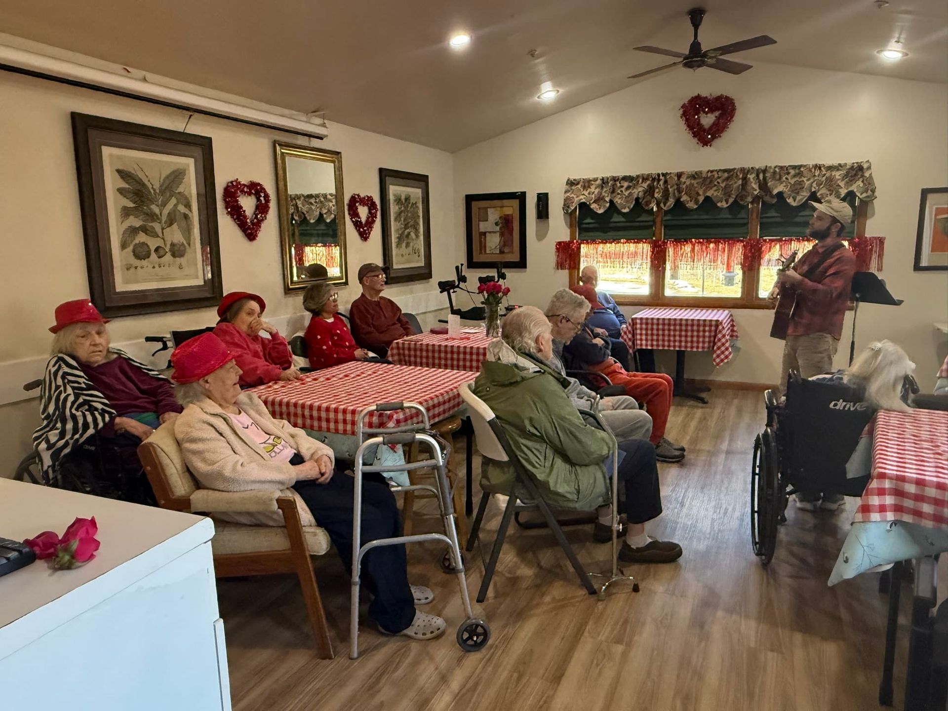 A group of people listens to a man playing guitar in a room decorated for Valentine's Day with red hearts and tablecloths.