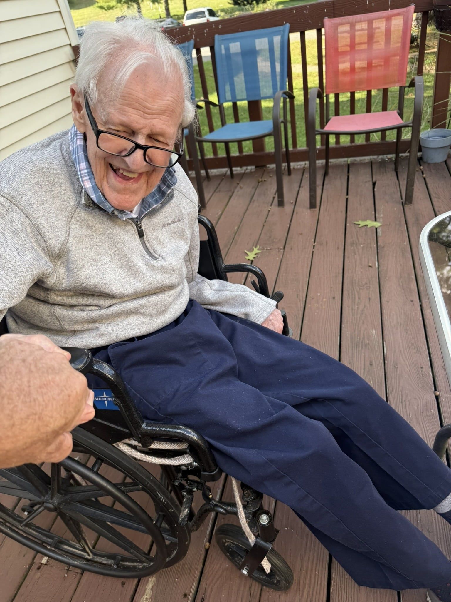 A smiling person in a wheelchair on a wooden deck with colorful chairs in the background.