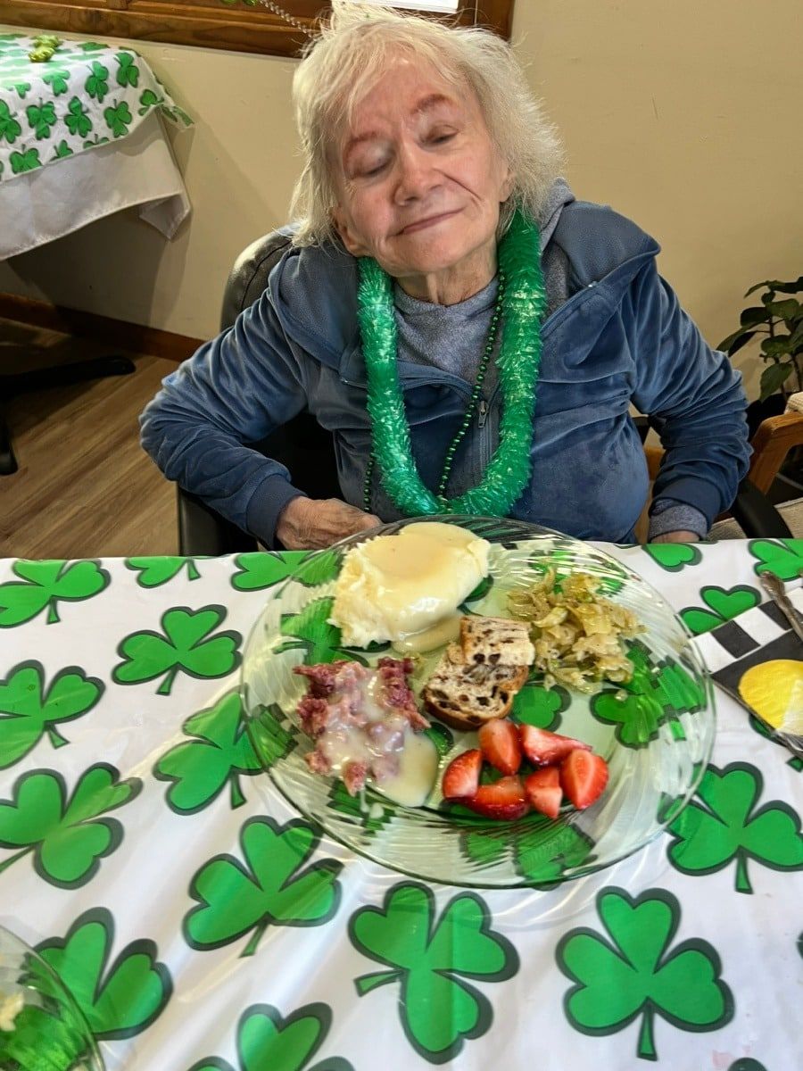 A smiling individual wearing a green lei sits at a table with a shamrock-patterned tablecloth, eating a plate of food.