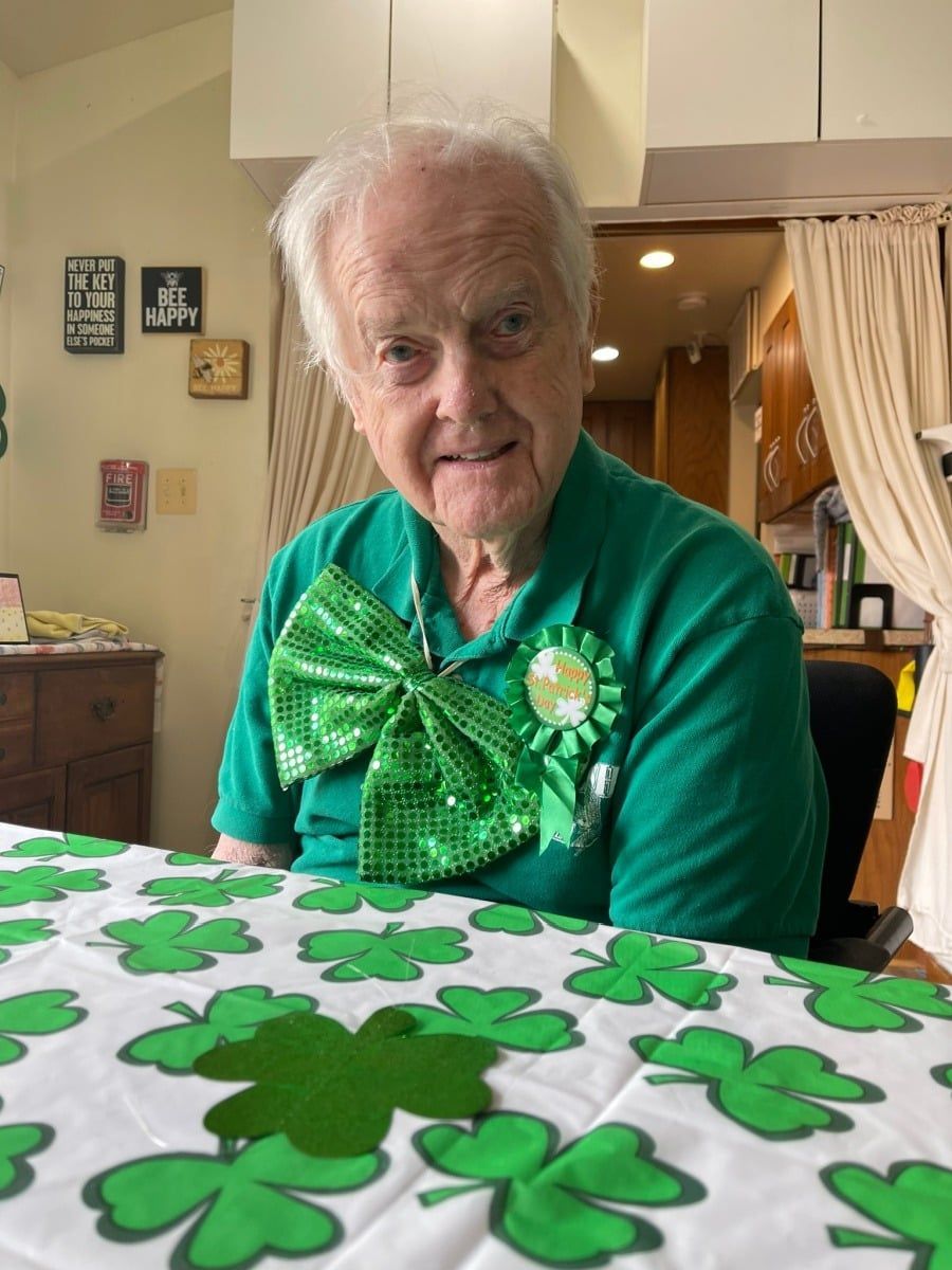 A smiling person wearing a green polo shirt, a large sequined bow, and a shamrock pin, seated at a clover-patterned table.