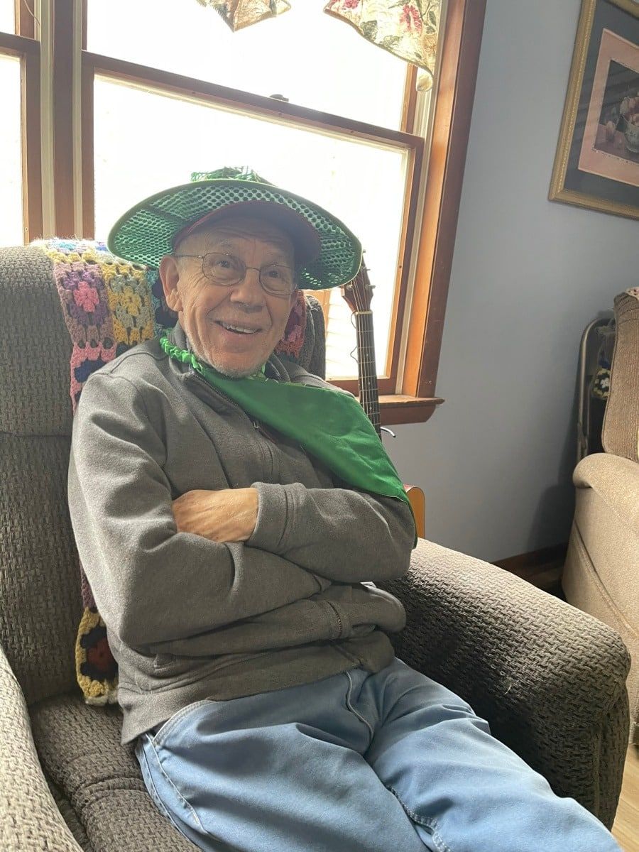 A person in a gray sweater and blue jeans sitting in a chair, wearing a green mesh hat and scarf, smiling.