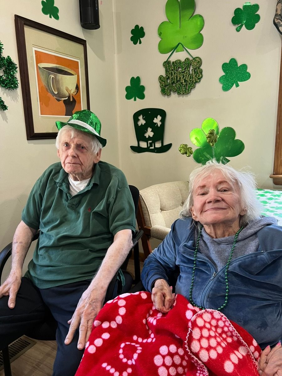 Two people sit together in a room decorated with shamrocks for St. Patrick's Day; both wear festive green hats and accessories.