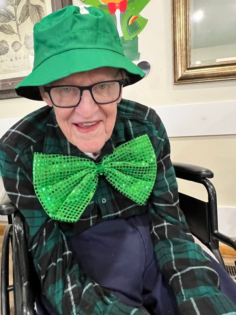 A person in a wheelchair wearing a green bucket hat, plaid shirt, and a large, sparkling green bow tie.