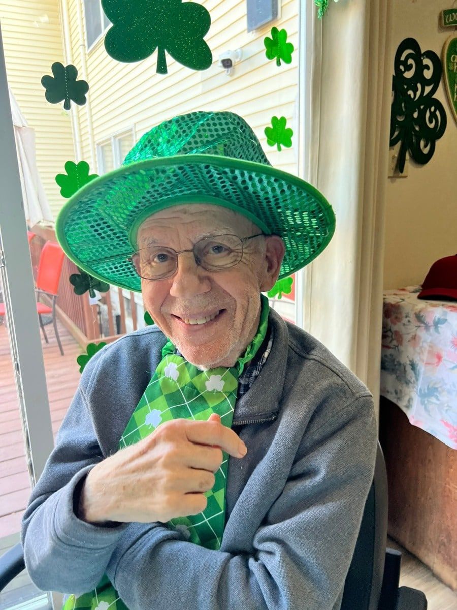 An individual smiles while wearing a bright green hat and a patterned green scarf, set against a background of shamrocks.