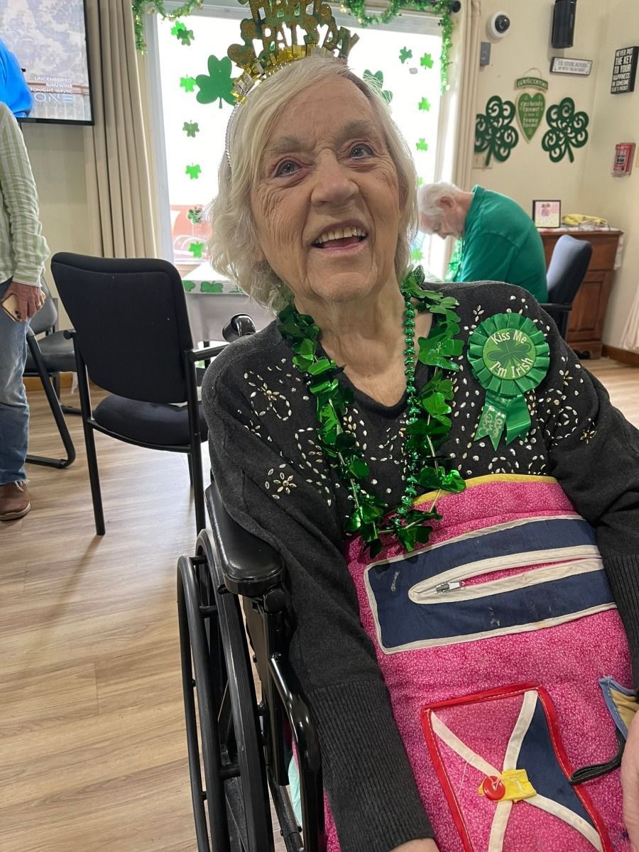 An individual in a wheelchair wears a green necklace and shamrock pin in a room decorated for St. Patrick’s Day.