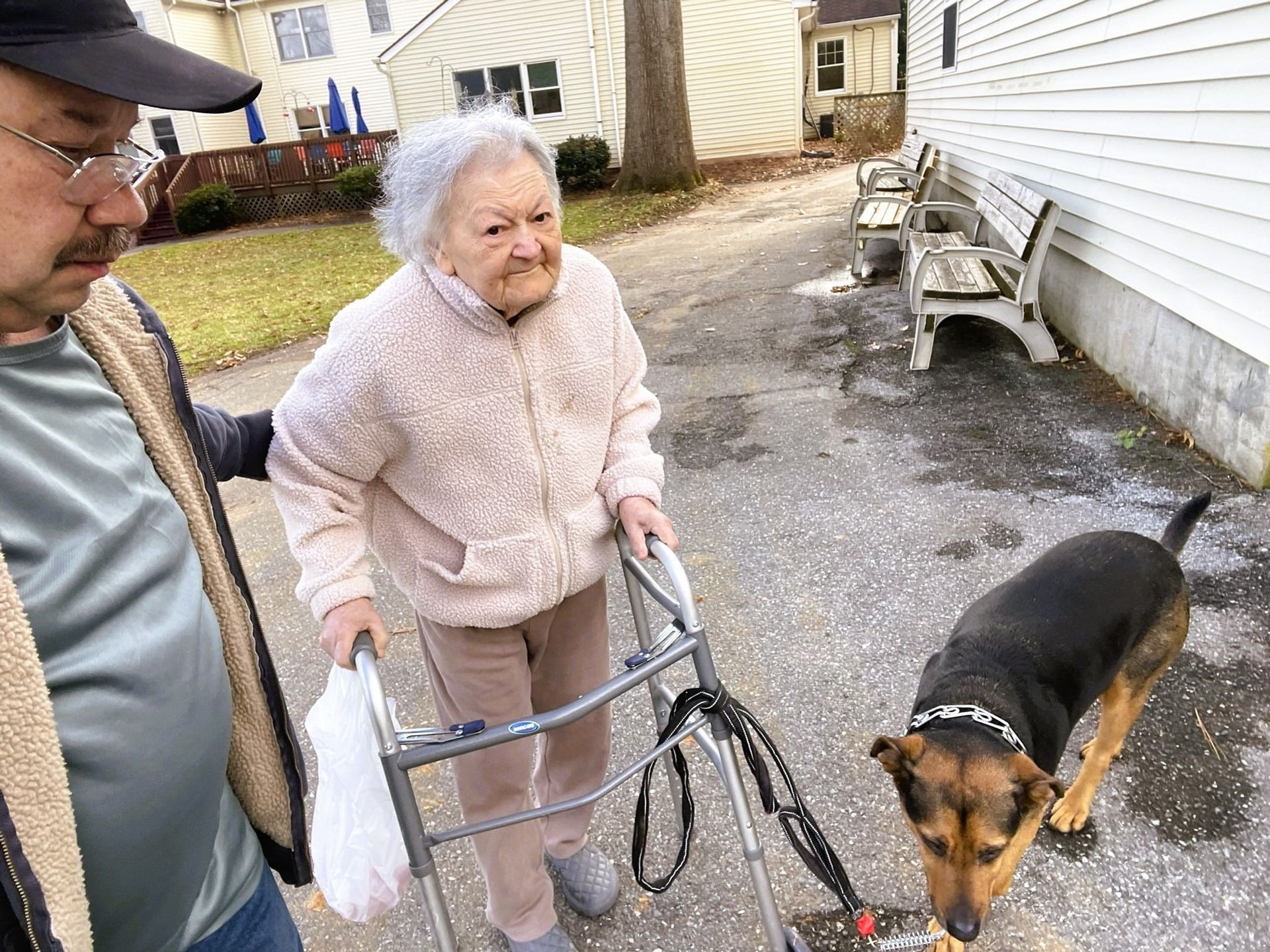 A person with a walker stands on a driveway with a companion and a dog on a leash near a building.