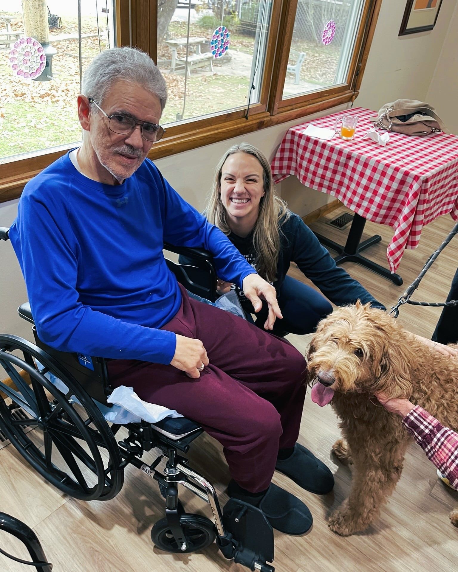 A smiling person in a wheelchair and a woman beside them pet a golden-brown dog in a room with a checkered table cloth.