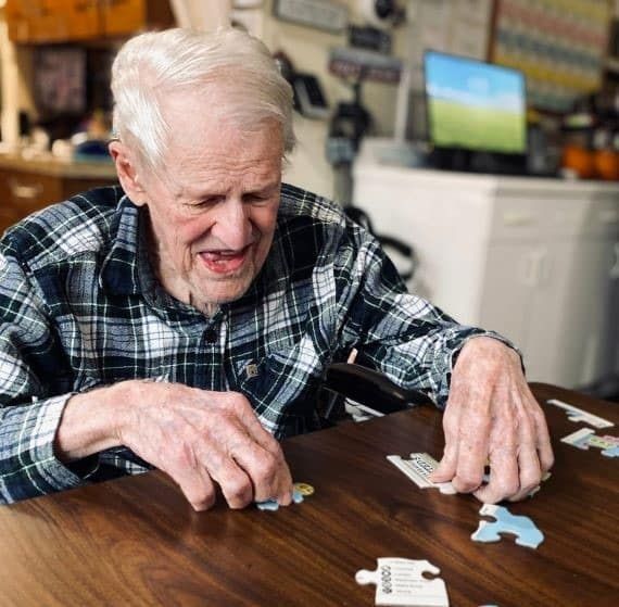 An older person in a plaid shirt sits at a wooden table, smiling while assembling pieces of a jigsaw puzzle.