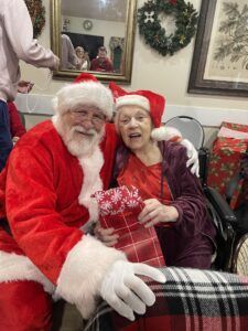 Santa Claus sits next to an individual in a holiday hat, both holding a wrapped gift in a decorated indoor setting.