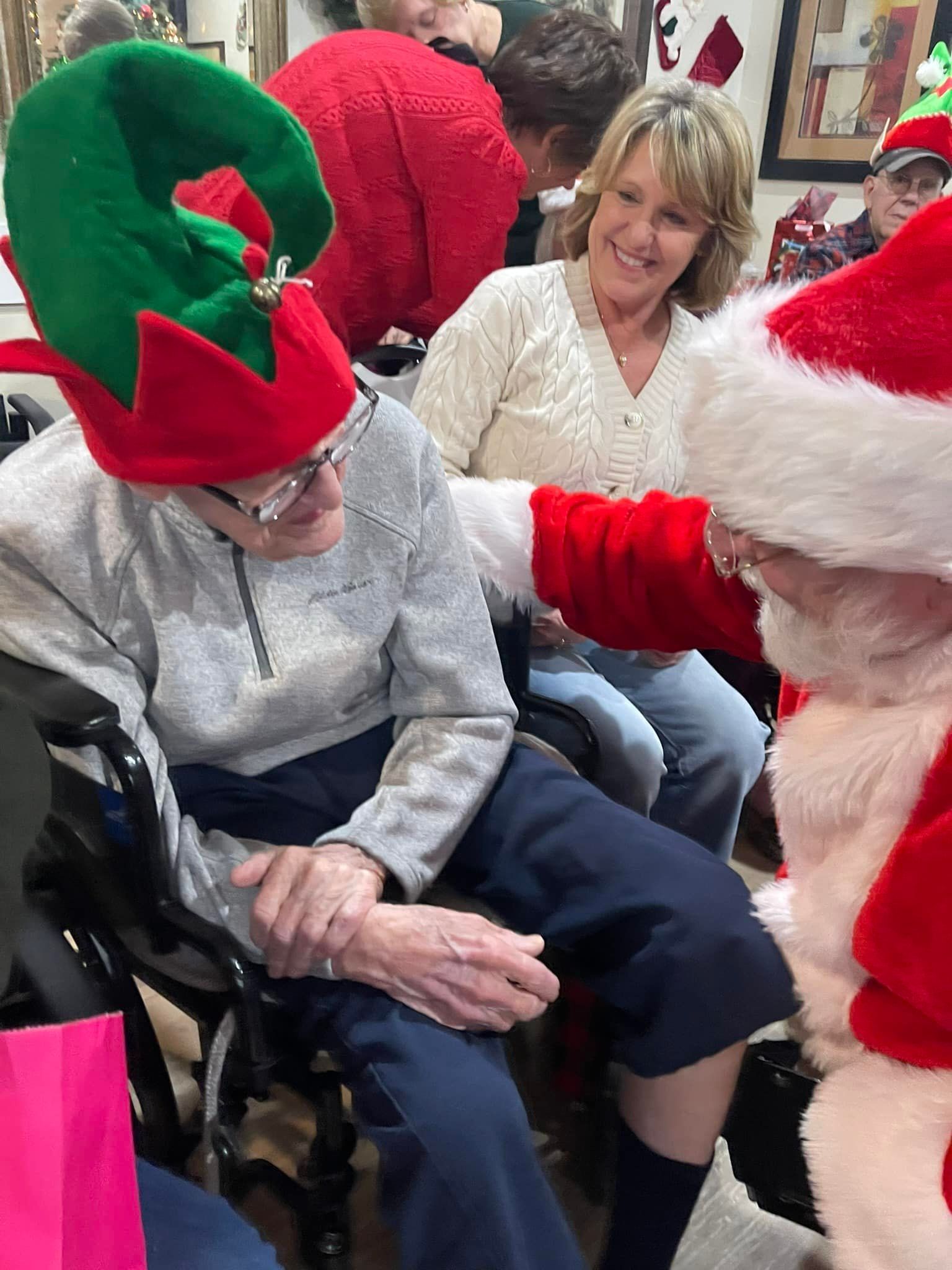 A person in an elf hat and wheelchair sits with a smiling woman while interacting with a person dressed as Santa Claus.