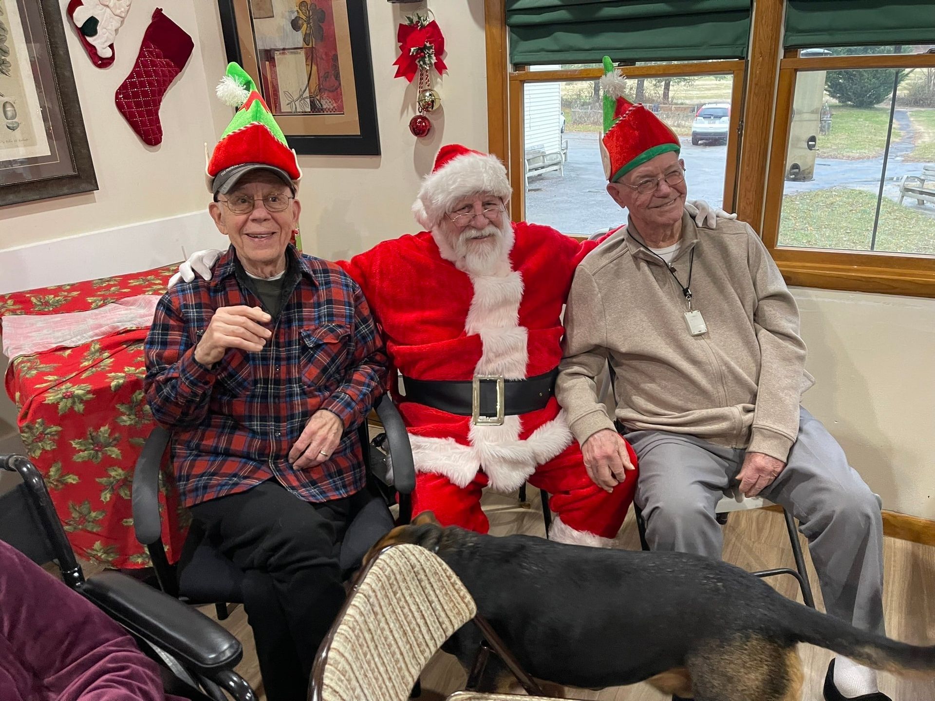 Three people, including one dressed as Santa Claus, wear elf hats in an indoor setting with a dog near the foreground.