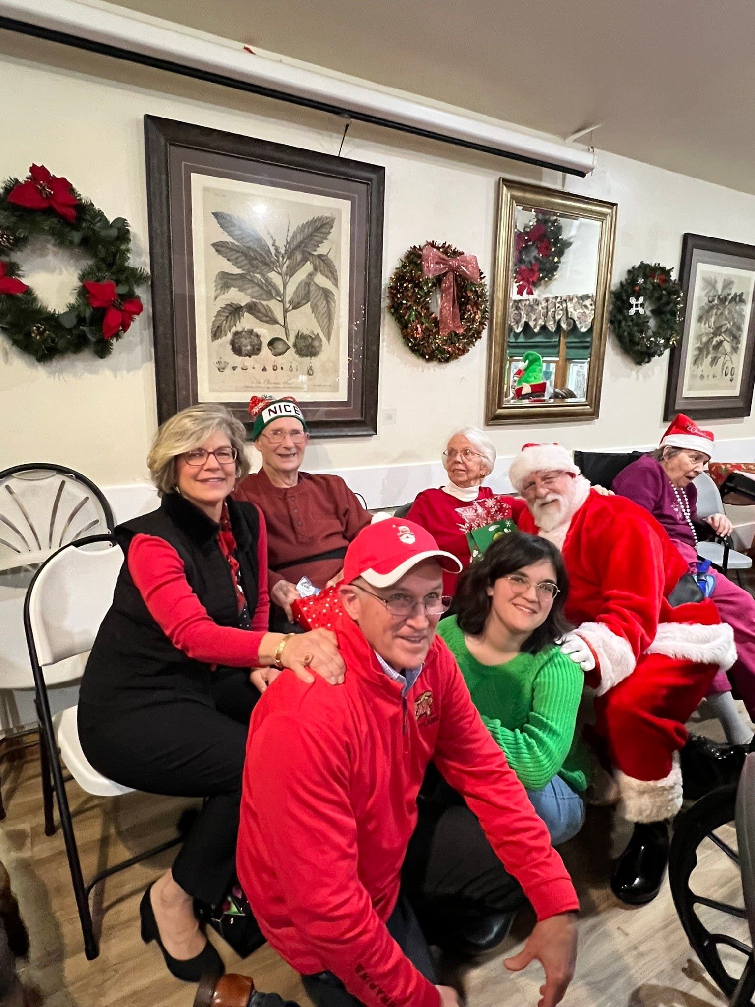 A group of people wearing holiday attire and Santa gear gather for a photo indoors, decorated with Christmas wreaths.