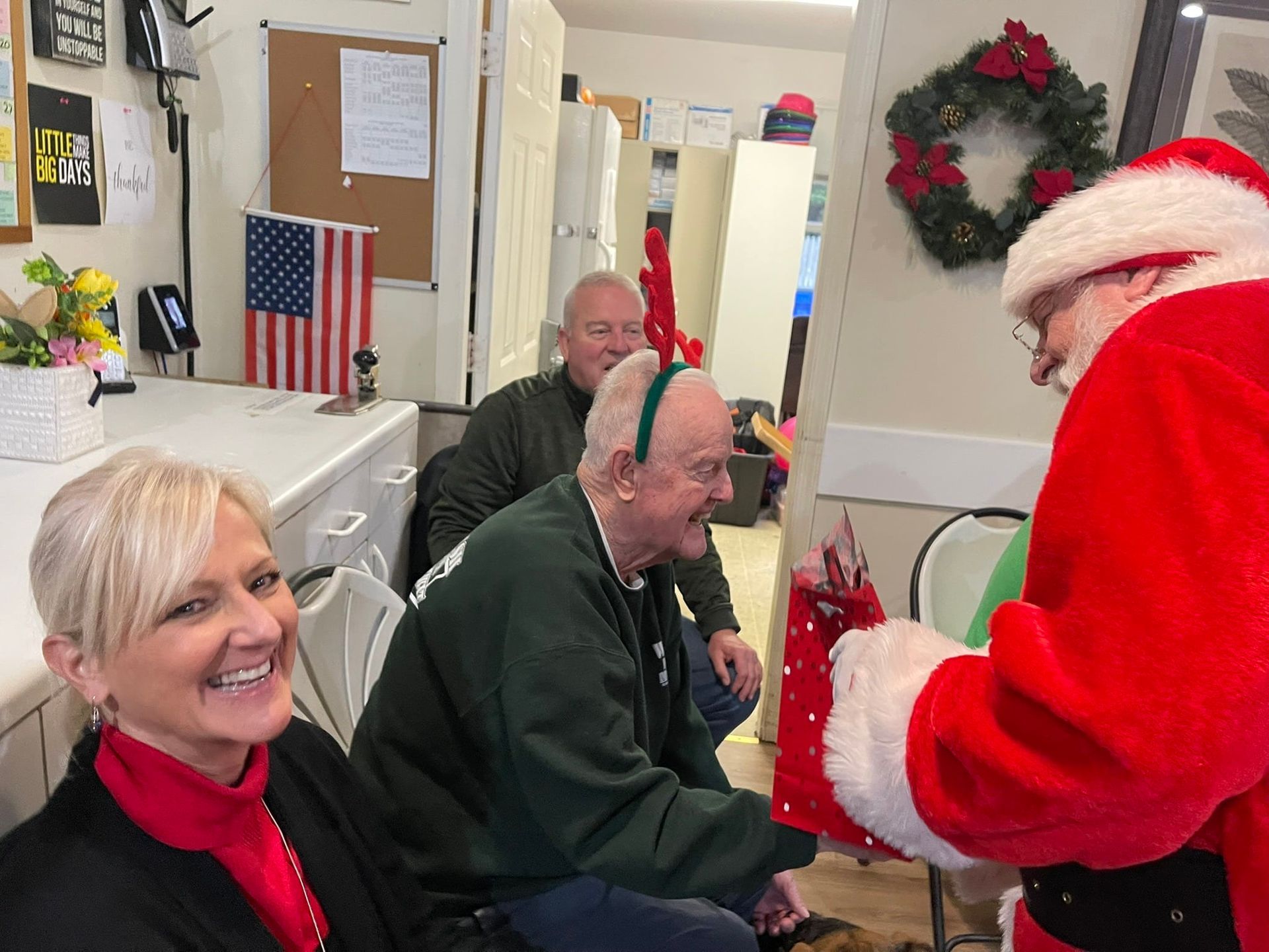Santa Claus hands a red gift bag to a man wearing a green headband while a smiling woman looks on in a festive room.