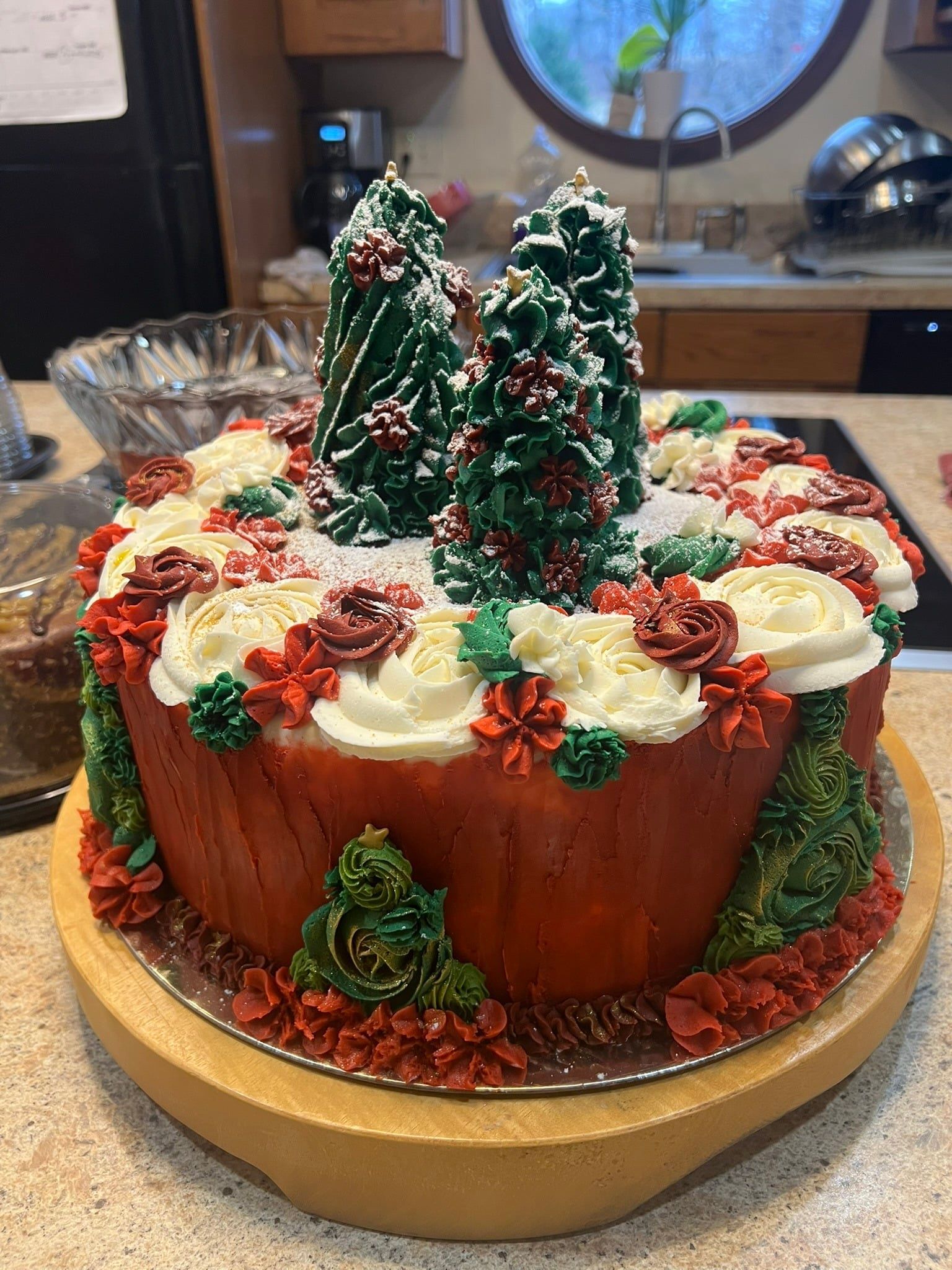 A holiday-themed cake on a wooden stand, topped with miniature frosted trees, red and white icing flowers, and sprinkles.