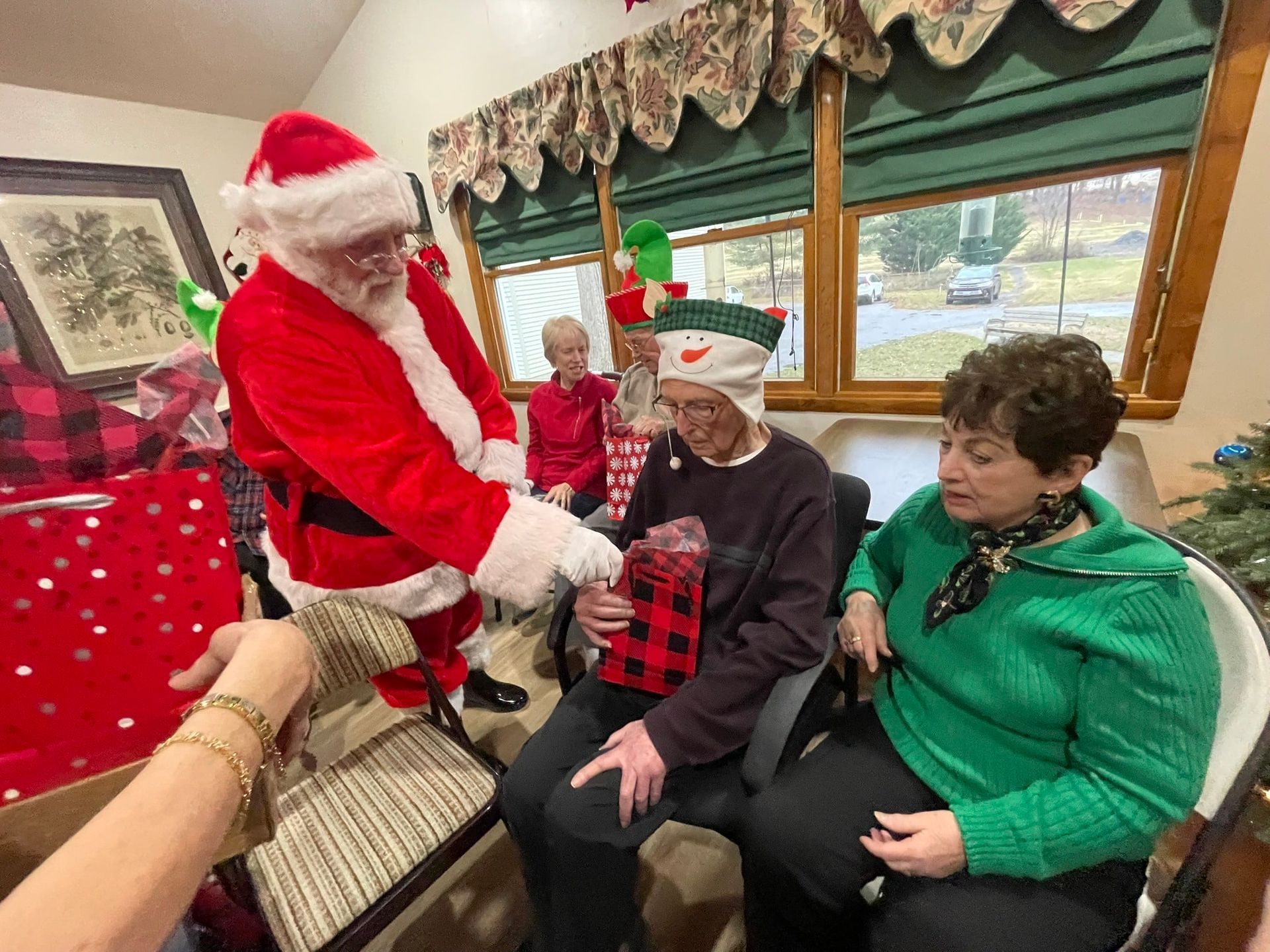 Santa Claus hands a wrapped gift to a person wearing a snowman hat while a woman in a green sweater looks on.