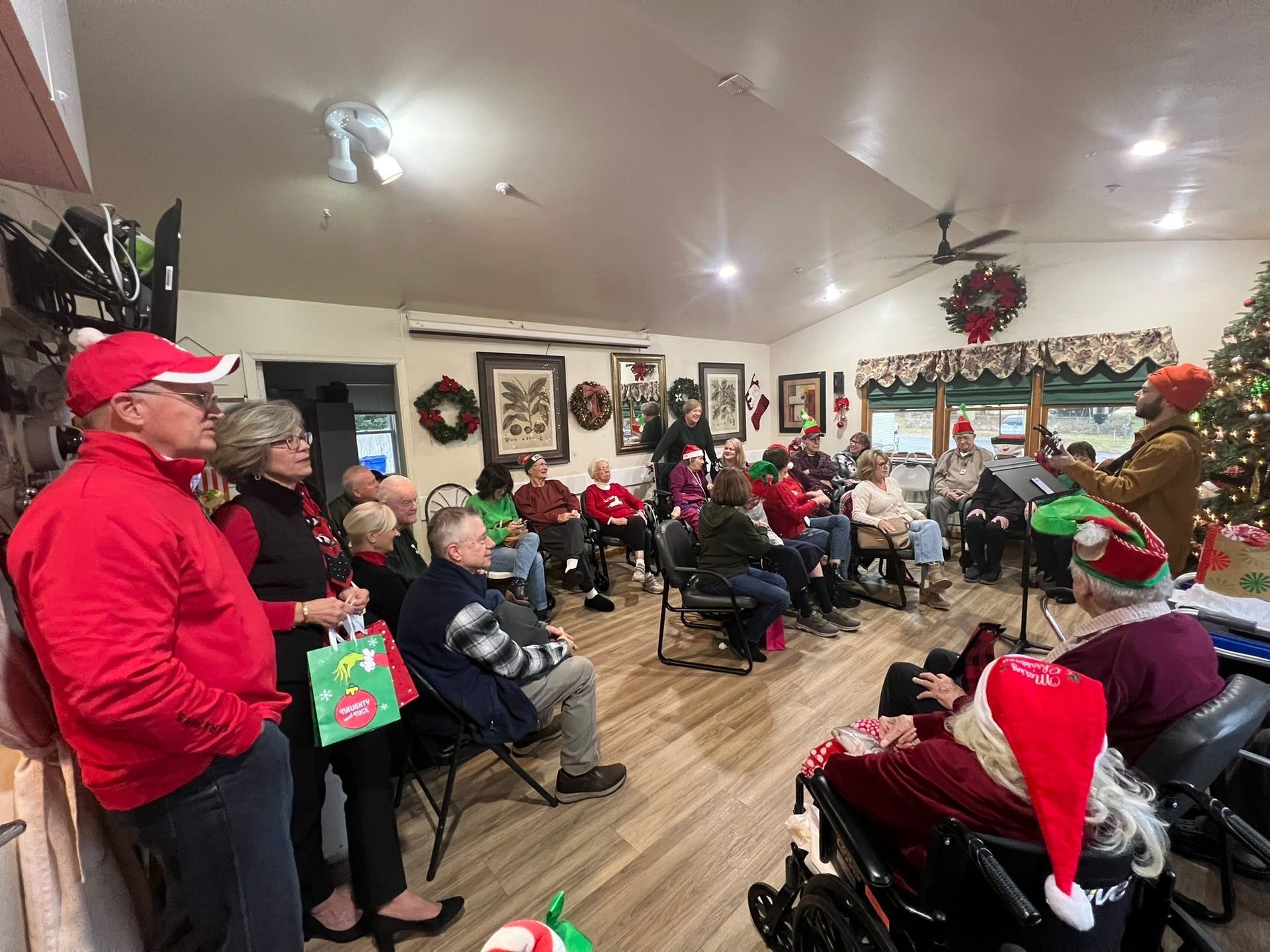 A group of people in a room decorated for Christmas, with one person playing guitar for the seated attendees.