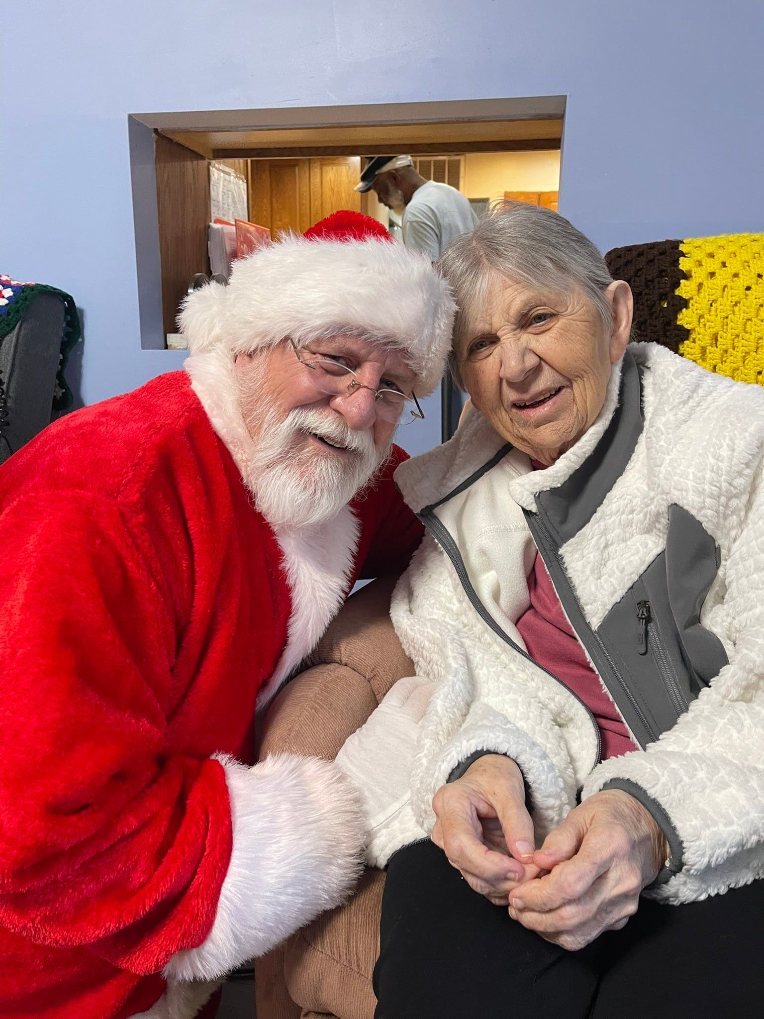 Santa Claus with his arm around a smiling person in a white fleece jacket, sitting together indoors.