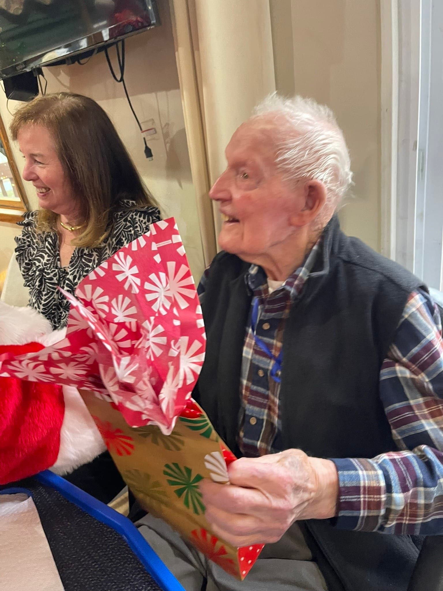 An older person in a plaid shirt and vest holds a gift bag with red tissue paper, smiling next to a woman in a black top.
