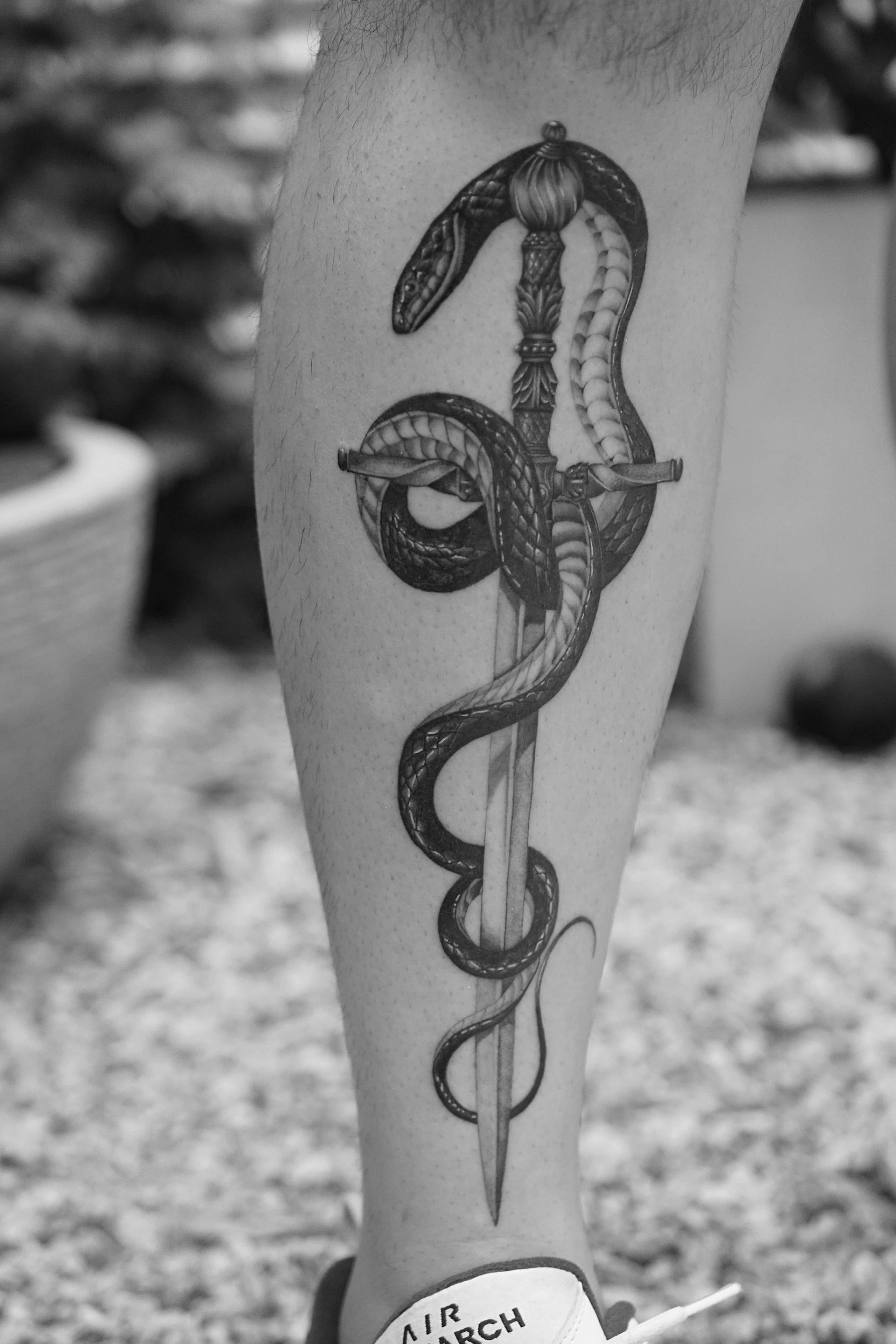 Black and white tattoo of a snake coiled around a sword on a person's calf.